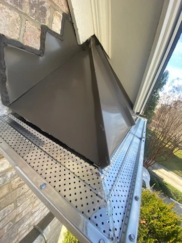 Perforated gutter guard mesh fitted below a downspout junction on brick